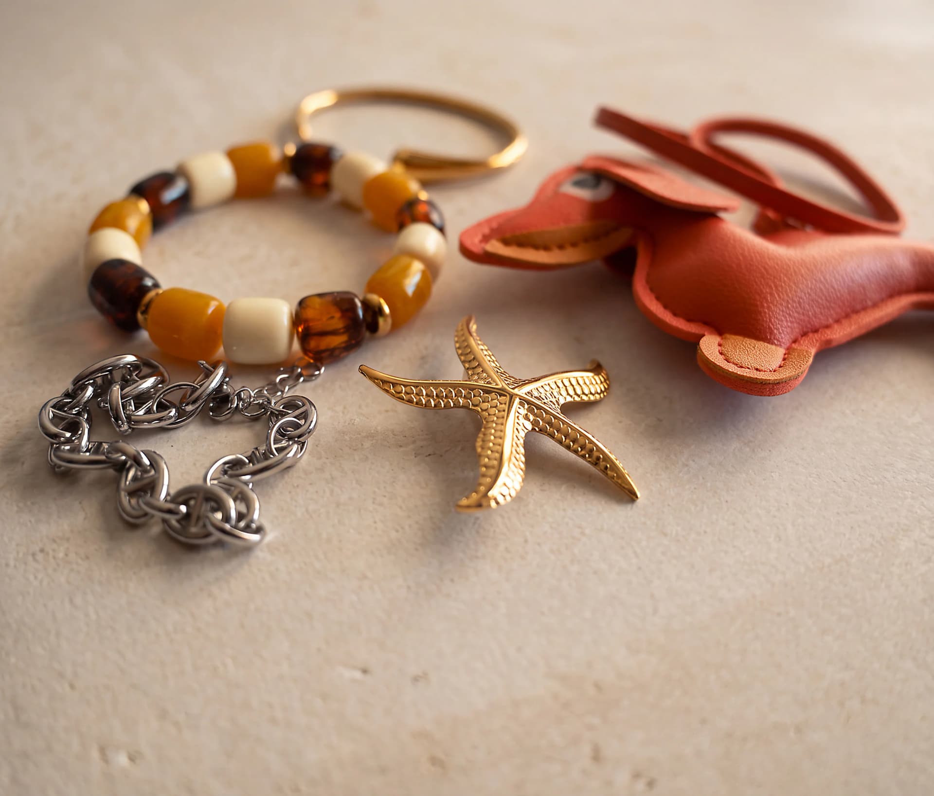 Um grupo de peças Synarava sobre uma superfície clara, incluindo pulseira de contas, bracelete dourado, pulseira de corrente prateada, pendente de estrela-do-mar dourada e pendente de mala em couro rosa com forma de pato