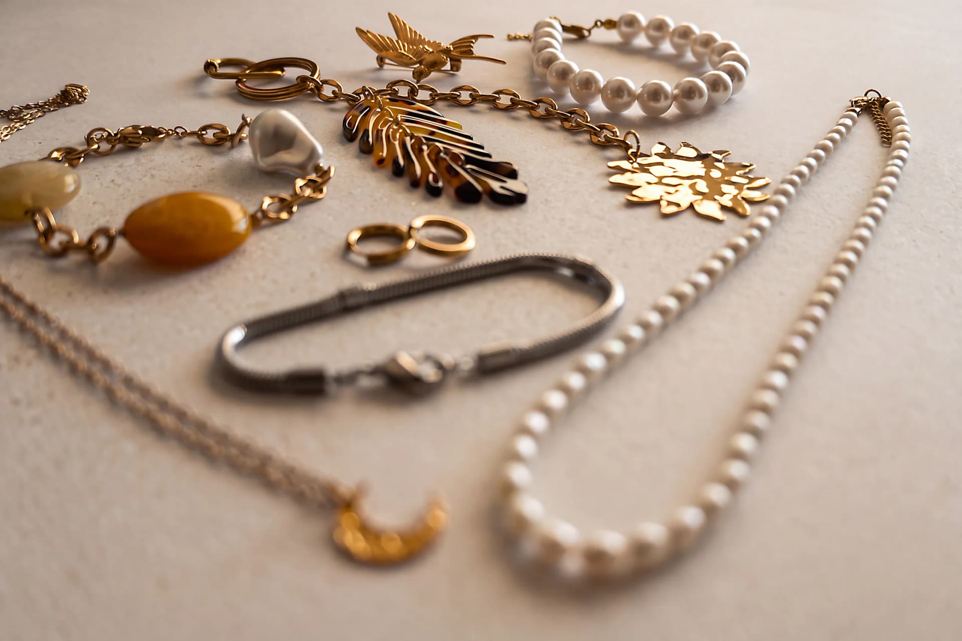 Flat lay of gold-tone jewellery on a pale stone surface: a swallow brooch, a pearl bracelet and necklace, a chain with a tortoiseshell leaf charm, a hammered sun pendant, amber beads, small hoops and a steel snake-chain bracelet.